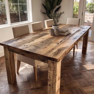 May include: A substantial wooden dining table with a reclaimed wood aesthetic. The table is roughly 183 cm long and 91 cm wide, supported by sturdy, square legs. Four upholstered chairs are arranged around the table. A plant and mugs are placed on the table.