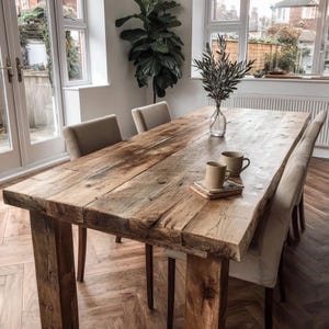 May include: A large, reclaimed wooden dining table with a natural finish. The table has a thick, rectangular top and sturdy, square legs. Two beige upholstered chairs are positioned on either side. The table is approximately 244 cm long.