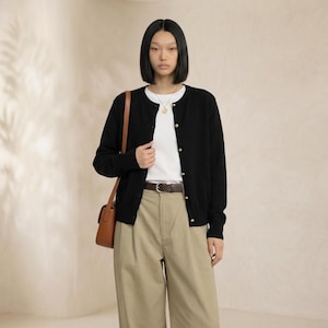 Op de afbeelding: Een persoon draagt een zwart vest met gouden knopen, een wit t-shirt en een kaki broek. Een bruine riem en een bruine schoudertas maken de outfit compleet. De achtergrond is neutraal.