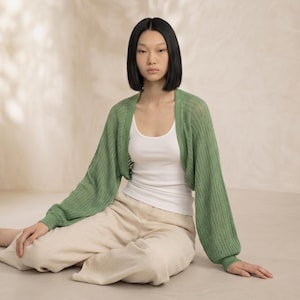 May include: A woman wearing a green, open-front bolero sweater with long sleeves. The sweater is paired with a white tank top and beige pants. The bolero has a ribbed knit texture and a relaxed fit, suitable for layering.