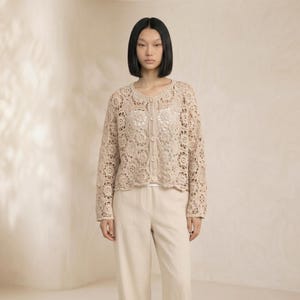 Puede incluir: Cárdigan de crochet beige con botones y mangas largas. El cárdigan presenta un delicado estampado floral y se combina con pantalones color crema. La modelo lleva un top blanco debajo del cárdigan.