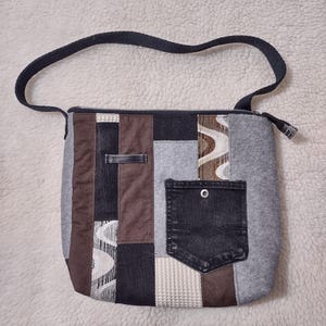 May include: A patchwork shoulder bag with a black strap and zipper closure. The bag features a variety of fabrics in shades of gray, brown, and black, including denim and patterned textiles. A black denim pocket is sewn onto the front.