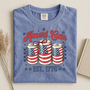 May include: A blue t-shirt featuring the text "Ameri Can" in red script above three cans decorated with the American flag. The text "EST. 1776" is below the cans, with stars on the shirt.
