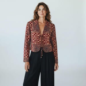 Puede incluir: Una blazer estampada en tonos rojos, beige y azul marino, combinada con pantalones negros de pernera ancha. La blazer presenta un diseño geométrico único y un cierre de lazo en la cintura, ofreciendo un look elegante y versátil.