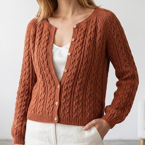 May include: A rust-colored, cable-knit cardigan with a round neckline and button-up closure. The long-sleeved sweater features a textured pattern and is paired with white pants. The buttons are a light, natural wood color.