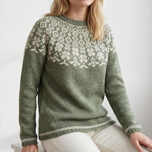 May include: A green sweater with a white geometric pattern across the upper chest and shoulders. The sweater has long sleeves and a ribbed hem and cuffs with a white and green design. The model is wearing white pants.