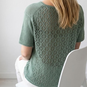 Pode incluir: Uma blusa de malha verde sálvia de manga curta com decote redondo e um padrão de trabalho aberto detalhado nas costas. A blusa é feita de um material macio e respirável. A modelo está usando calças brancas.