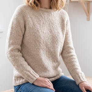 以下が含まれることがあります： ベージュ色のニットセーター。長袖とクルーネックのデザイン。セーターは質感があり、ゆったりとしたフィット感。袖はラグランスリーブスタイル。柔らかく暖かい素材で作られています。
