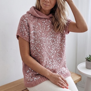 May include: A dusty rose-colored, short-sleeved sweater with a hood, heavily embellished with scattered, shiny sequins. The sweater is knitted and has a relaxed fit. The model is wearing white pants.