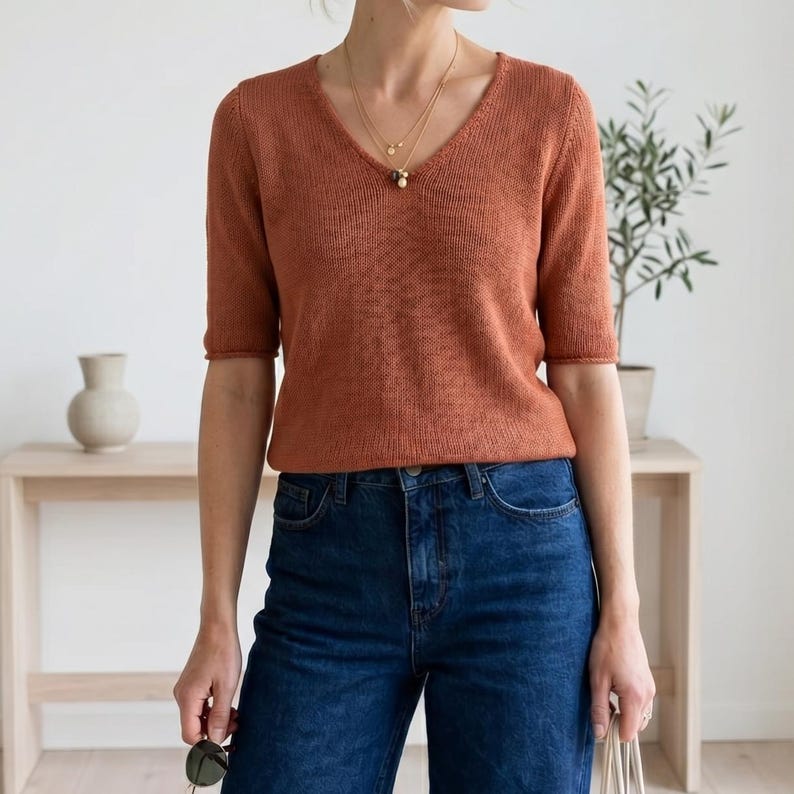 Puede incluir: Un su&eacute;ter de cuello en V de color &oacute;xido y manga corta. El su&eacute;ter se combina con vaqueros de mezclilla azul oscuro. Se lleva un collar de oro. El fondo incluye una mesa de madera, un jarr&oacute;n y una planta en maceta.
