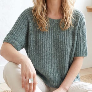 May include: A short-sleeved, ribbed knit top in a muted teal color. The top has a round neckline and a slightly boxy fit. The knit fabric has a subtle texture and a speckled appearance. The model is wearing the top with white pants.