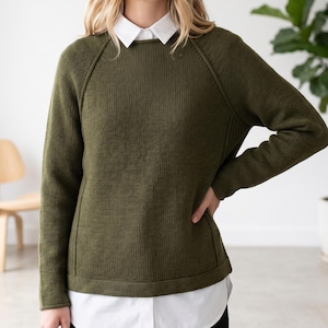 Op de afbeelding: Olijfgroene trui met een ronde hals en lange mouwen. De trui heeft een geribbelde textuur en een wit overhemd met kraag eronder. De mouwen hebben een raglan ontwerp. De trui is effen van kleur en lijkt gemaakt van een zacht gebreid materiaal.