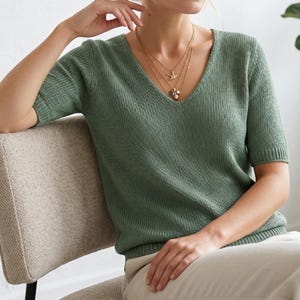 May include: A sage green V-neck knit top with short sleeves. The top is worn with layered gold necklaces. The person is seated on a beige chair. The top is made of a soft, breathable material.