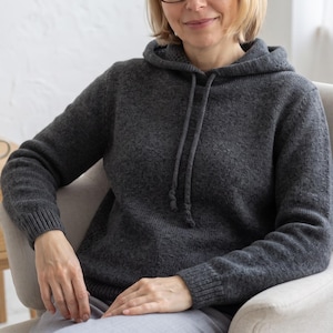May include: A dark charcoal grey knit hoodie featuring long sleeves with ribbed cuffs, a matching ribbed hem, and a hood with two integrated drawstrings. The garment is crafted from a textured knit fabric with a consistent dark grey colour throughout.