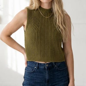 May include: Olive green sleeveless sweater with a ribbed knit design. The sweater has a mock neck and a cropped length. A gold chain necklace is worn with the sweater. The model is wearing blue jeans.