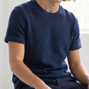 Könnte beinhalten: Ein kurzärmeliges, marineblaues T-Shirt mit einer strukturierten, gewebten Stoffoberfläche. Das Kleidungsstück verfügt über einen klassischen Rundhalsausschnitt sowie gerippte Details am Kragen und an den Ärmelbündchen für einen strukturierten Look.