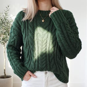 Può includere: Un maglione verde scuro a trecce con scollo tondo. Il maglione ha un design spesso e strutturato e maniche lunghe. È visibile una collana con pendente a cuore dorato. La persona indossa jeans bianchi. È un capo caldo e accogliente.