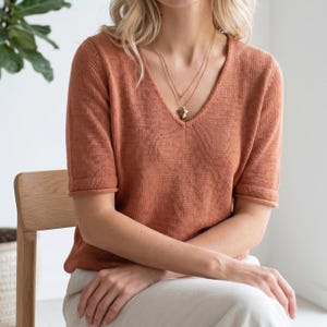 Puede incluir: Una camiseta de punto de color &oacute;xido con mangas cortas y cuello en V. La camiseta se combina con un collar dorado. La modelo est&aacute; sentada en una silla de madera. La camiseta tiene un dise&ntilde;o sencillo y cl&aacute;sico.