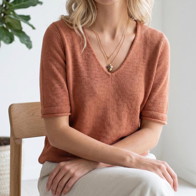 Puede incluir: Una camiseta de punto de color &oacute;xido con mangas cortas y cuello en V. La camiseta se combina con un collar dorado. La modelo est&aacute; sentada en una silla de madera. La camiseta tiene un dise&ntilde;o sencillo y cl&aacute;sico.