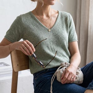 Puede incluir: Una camiseta verde claro con cuello en V y manga corta. La camiseta se combina con vaqueros azules. La persona sostiene gafas con estampado de carey y una peque&ntilde;a bolsa con cord&oacute;n. La persona est&aacute; sentada en una silla de madera.