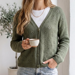 Pode incluir: Cardigã verde-oliva com decote em V e fecho de botões. O suéter de malha apresenta um padrão de losangos e é combinado com uma camiseta branca e um colar dourado. Uma pessoa segura uma caneca de cerâmica.