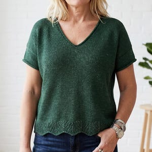 May include: A dark green, short-sleeved knitted top with a V-neck. The top has a textured appearance and a scalloped hemline. The model is wearing the top with blue jeans. The top is a comfortable and stylish garment.