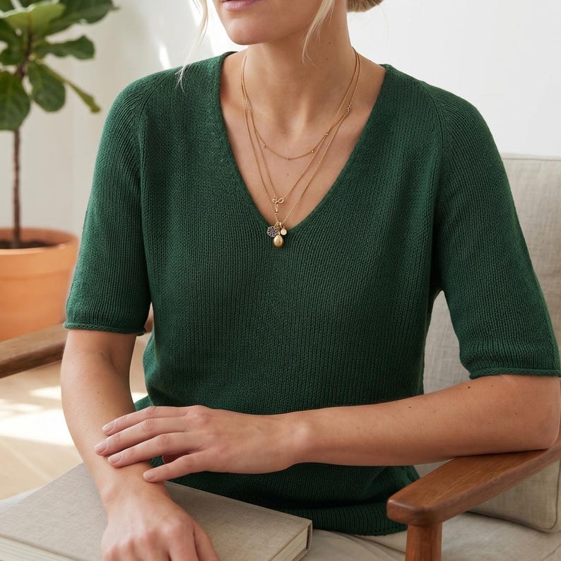 1. Woman wearing a green knitted V-neck short sleeve top with layered necklaces  
2. Close-up of lightweight knit tee showing smooth stockinette texture and neckline  
3. Minimalist summer knit top styled with neutral pants and jewelry  
4. Detail of V-neck shaping on handmade knitted short sleeve sweater  
5. Soft lightweight knitted tee worn casually indoors with modern styling