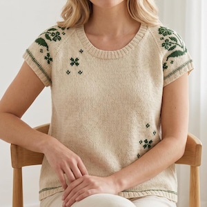 Può includere: Un top in maglia color crema con maniche corte e scollo tondo. Il top presenta motivi floreali verdi sulle spalle e sparsi sul davanti. I bordini verdi accentuano le maniche e lo scollo. La persona è seduta su una sedia di legno.