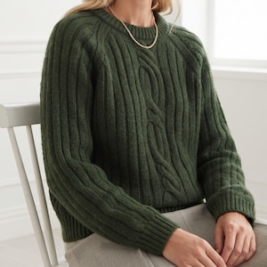 May include: A dark forest green long-sleeve sweater featuring a chunky cable knit pattern on the front and ribbed knit detailing on the sleeves and sides. The sweater has a round crew neckline and is paired with a simple gold chain necklace.