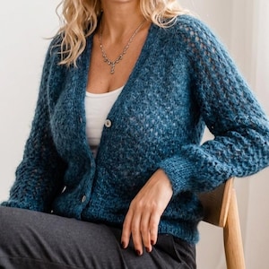 May include: A teal blue cardigan with a V-neck and button closure. The knitted sweater has a textured pattern and long sleeves. The sleeves have a looser, open knit design. The cardigan is worn over a white top.