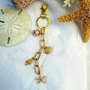 Könnte beinhalten: Goldfarbener Schlüsselanhänger mit einer Kette mit Muschel-, Seestern- und Blumenanhängern. Der Schlüsselanhänger hat einen Karabinerverschluss und einen großen Ring. Die Anhänger sind detailliert und fügen ein Küstenmotiv hinzu.
