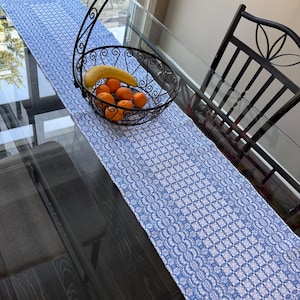 May include: A long rectangular table runner featuring a blue and white woven geometric pattern. A black metal wire fruit basket with a curved handle sits on the runner, containing one yellow banana and several small round orange fruits.