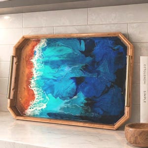May include: A rectangular wooden serving tray featuring a resin art design that mimics an ocean shoreline. The surface displays swirling shades of deep blue, turquoise, and white foam, transitioning into a sandy orange beach area with gold handles.
