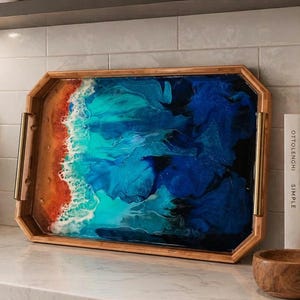 May include: A rectangular wooden serving tray with gold-toned metal handles. The interior surface features a resin art design depicting a beach scene with a sandy orange shore, white foam waves, and deep blue and turquoise ocean water.