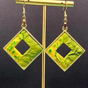 May include: A pair of gold-toned earrings with a square, diamond-shaped design. The earrings feature a yellow and green marbled pattern within a gold frame, with a square cut-out in the center. The earrings are displayed on a gold stand.