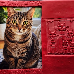 May include: A rectangular red frame featuring embossed cat silhouettes along the border and a raised relief panel on the right side depicting five stylized cats. The frame surrounds a photograph of a brown tabby cat with green eyes looking forward.