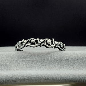 May include: A silver-toned metal ring featuring a continuous, raised relief pattern of swirling, vine-like motifs. The design has a darkened, oxidized finish in the recessed areas to highlight the intricate, curved shapes along the band.