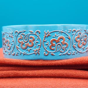 May include: A turquoise blue, cylindrical bracelet with an ornate, repeating floral and scroll design in a contrasting copper colour. The bracelet rests on folded orange fabric, against a bright blue background.