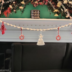 May include: A macrame garland featuring a twisted off-white cord base adorned with hanging ornaments. The decorations include two red knotted trees, two red and white striped circular wreaths, and a central off-white macrame angel figure.