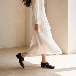 May include: Black leather shoes with a square toe and a strap design. The shoes have a low heel and are worn with a long white dress. The image is taken in a room with neutral-colored walls and flooring.