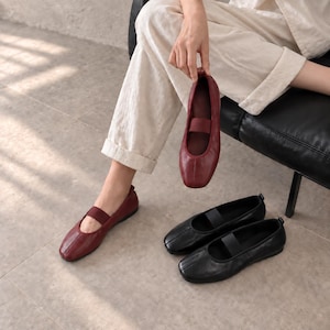 May include: A pair of dark red leather ballet flats with a square toe and a matching elastic strap across the instep, alongside a pair of black leather ballet flats featuring the same square toe design and a black elastic strap across the instep.