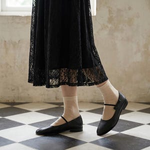 May include: A black lace skirt paired with sheer beige ankle socks and black leather Mary Jane flats. The shoes feature a thin strap across the instep and a low heel. The skirt has a floral lace pattern and falls below the knee.