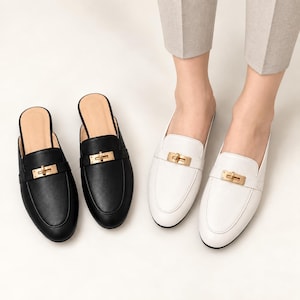 May include: A pair of black backless loafers and a pair of white backless loafers, each featuring a horizontal strap across the vamp with a gold-toned metal turn-lock closure. The shoes have a rounded toe design and a smooth, solid-colored finish.