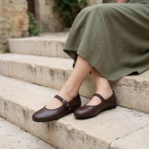 May include: A pair of dark brown leather Mary Jane shoes with a rounded toe, a thin strap across the instep, and a small silver-toned metal buckle. The shoes feature a flat sole and a smooth finish, worn with a long olive green skirt.