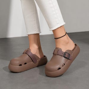 May include: Brown clogs with a strap and buckle detail. The clogs have a rounded toe and small ventilation holes. The shoes are worn with white pants. The clogs are a neutral color and appear to be made of a rubber-like material.