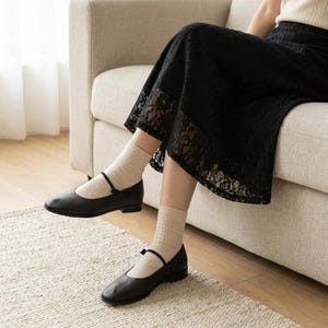 May include: A black lace midi skirt paired with beige ribbed crew socks and black leather Mary Jane flats featuring a thin strap across the instep. The skirt has a scalloped hemline and a floral pattern, while the shoes have a low block heel.