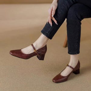 May include: Dark brown Mary Jane shoes with a square toe, a thin adjustable strap across the instep featuring a small gold-toned buckle, and a chunky, geometric block heel. The shoes are worn with dark charcoal grey denim jeans.