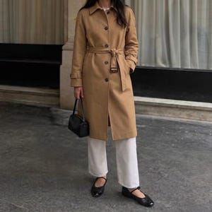 May include: A tan trench coat with a collared neckline, long sleeves, and a matching waist belt with a buckle. The coat features a button-down front closure. A small black handbag is held at the side, paired with white trousers and black flats.