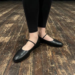 May include: A pair of black leather ballet flats featuring a rounded toe, a low heel, and a thin, delicate strap across the instep. The shoes have a smooth, matte finish and are worn with solid black leggings, showcasing a simple and functional design.