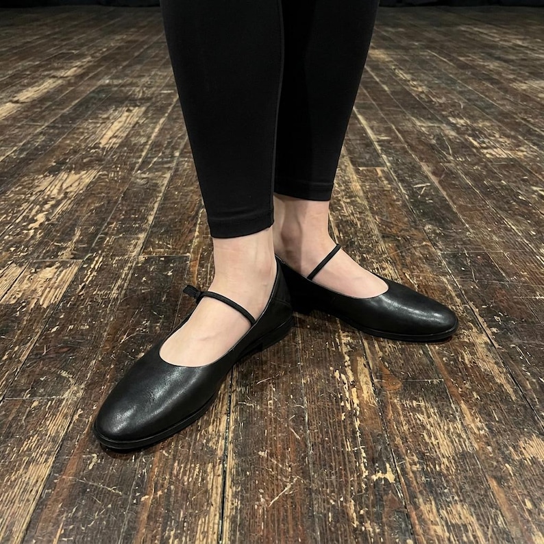 May include: A pair of black leather ballet flats featuring a rounded toe, a low heel, and a thin, delicate strap across the instep. The shoes have a smooth, matte finish and are worn with solid black leggings, showcasing a simple and functional design.
